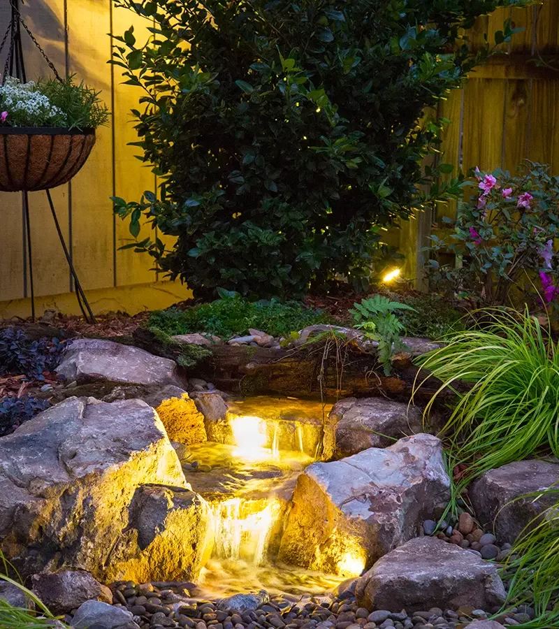 Evening garden waterfall with soft lighting, stones, and lush plants creating a tranquil atmosphere